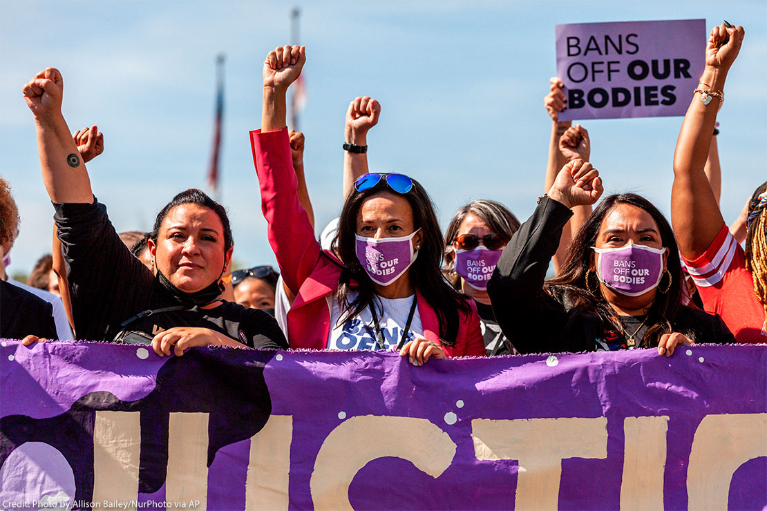 Three women with raised fists, wearing masks with "BAN OFF OUR BODIES" printed on them and carrying a purple banner, lead other protestors at the Women's March Rally For Abortion Justice In Washington, D.C.