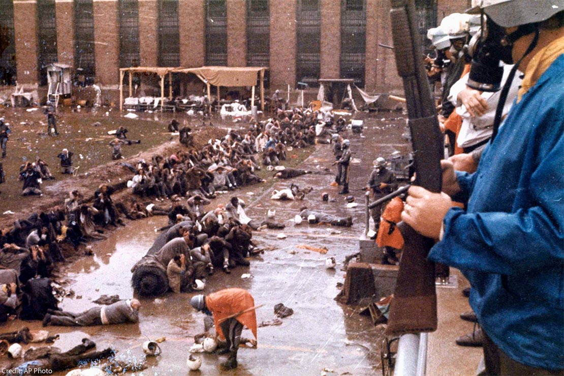 With helmets, gas masks and armed with shotguns, state police overlook the Attica prison yard which is filled with prisoners, face down in mud with their hands behind their heads. Other policemen are in orange ponchos are walking and searching the...