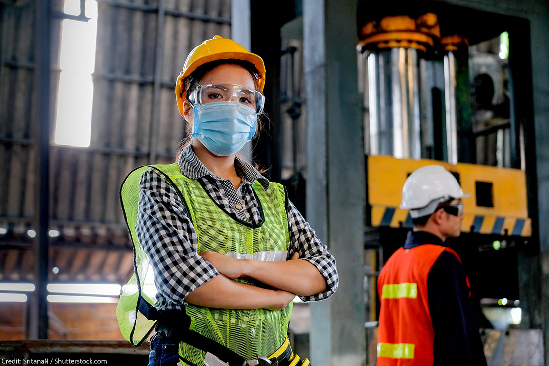 A technician wearing a surgical mask and hard hat stands with arms crossed in a factory.