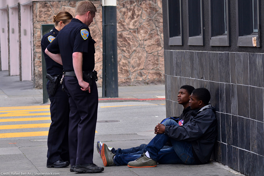 Two San Francisco police officers interrogating and standing over two Black American men seated on the sidewalk. Overall, Black Americans are arrested at 2.6 times the per-capita rate of all other Americans.