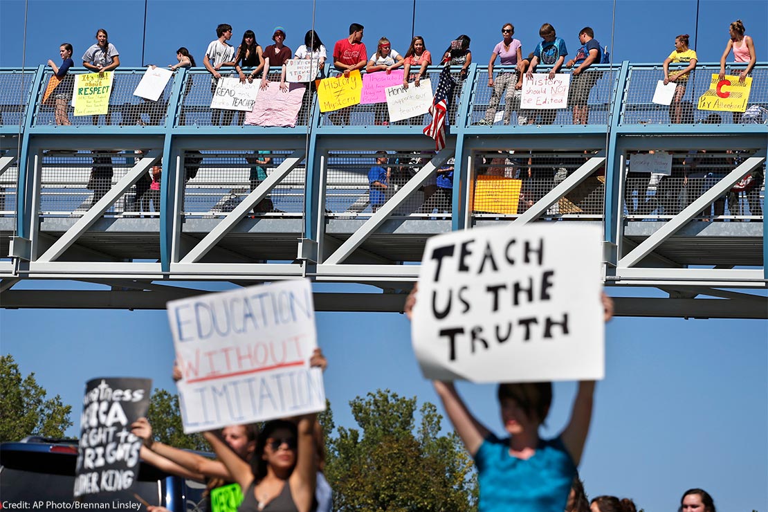 In the foreground (at an intersection,) two protesters carry signs with one reading "EDUCATION WITHOUT LIMITATION" and the other "TEACH US THE TRUTH", while in the background, other student demonstrators line an overpass protesting a proposal to...