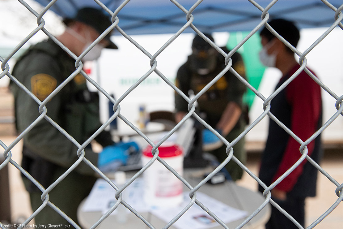 CBP agents wearing PPE processing someone wearing a mask behind a chain-link fence.