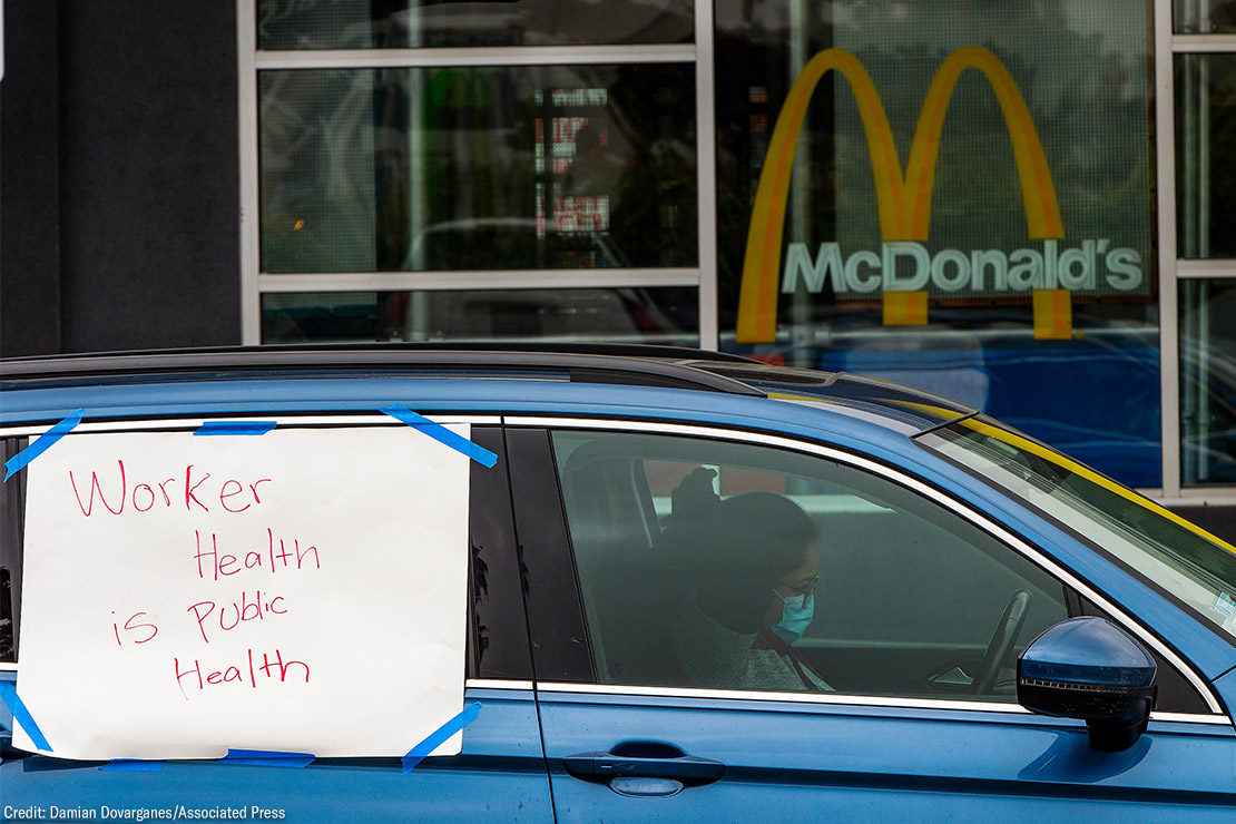 A protester in a car with a sign reading "Worker Health is Public Health" at a socially distanced protest.