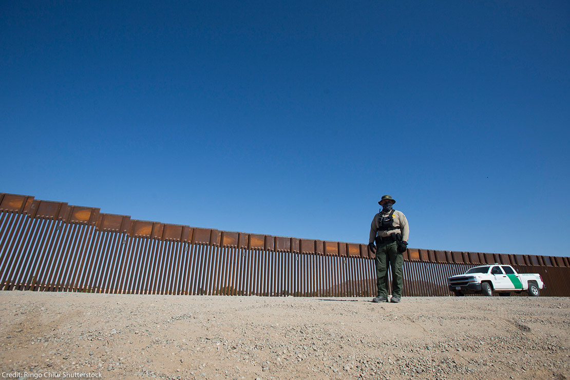 A uniformed individual in front of a portion of U.S., Mexico border wall.