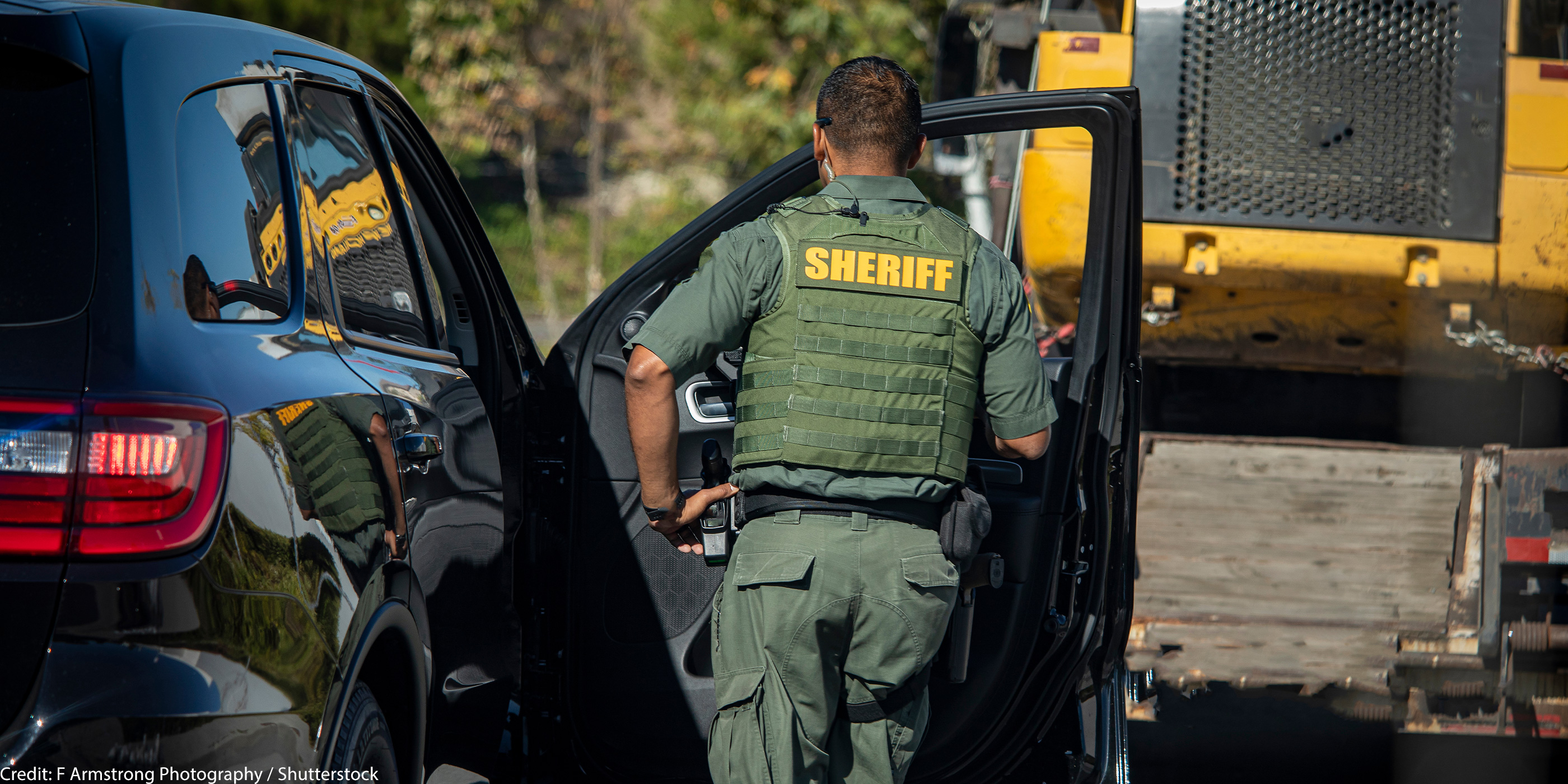 Shot of sheriff (wearing a bulletproof vest) from behind walking towards the open door of police car.