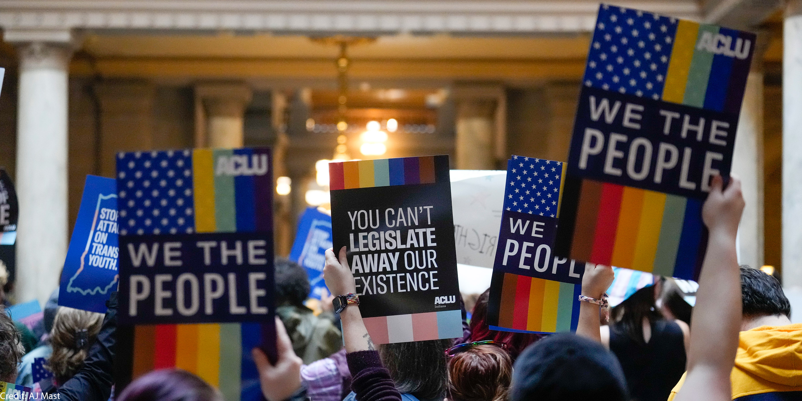 Individuals in a group holding ACLU-branded signs saying "We the People," and "You Can't Legislate Away Our Existence."