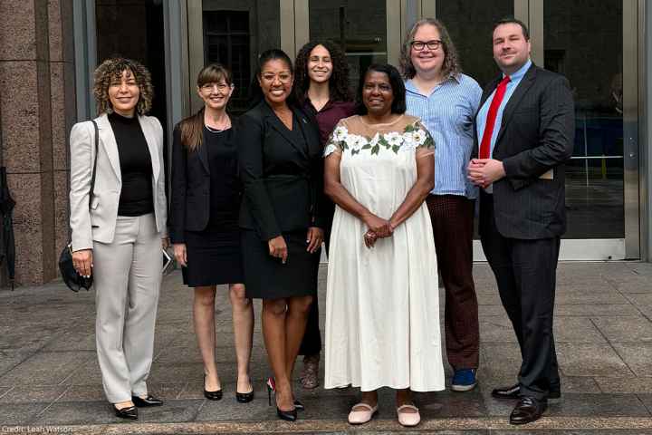 A smiling Leah Watson (a black woman wearing glasses, black business attire and a pin on her left shoulder stands next to a woman wearing a white flower dress wearing glasses and black) gathers for a picture with colleagues.