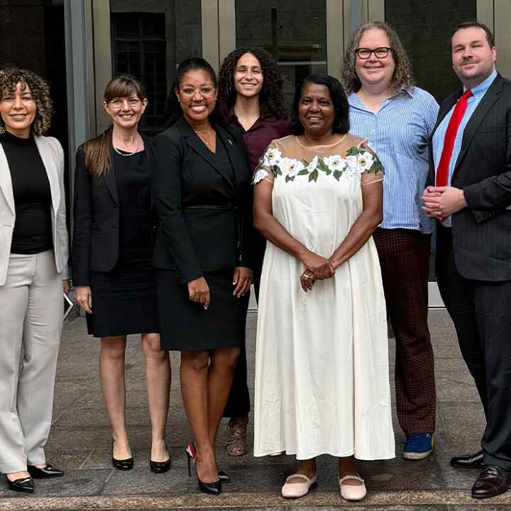 A smiling Leah Watson (a black woman wearing glasses, black business attire and a pin on her left shoulder stands next to a woman wearing a white flower dress wearing glasses and black) gathers for a picture with colleagues.