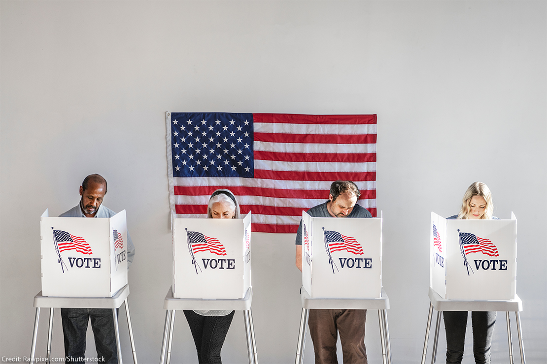 With and American flag in the background, four people in the act of voting, stand behind voter booths as they make their selections.